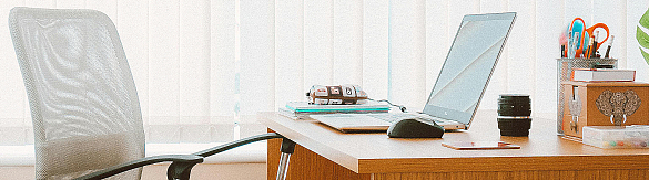 Ausschnitt eines hölzernen Schreibtisches mit Computer darauf. Davor steht ein moderner Bürostuhl