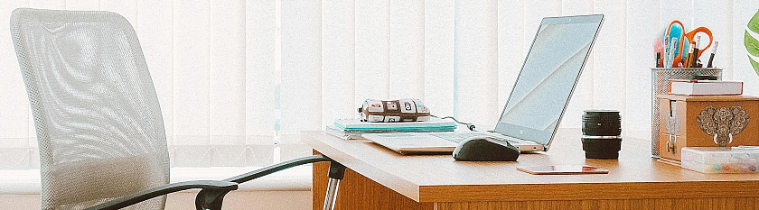 Ausschnitt eines hölzernen Schreibtisches mit Computer darauf. Davor steht ein moderner Bürostuhl